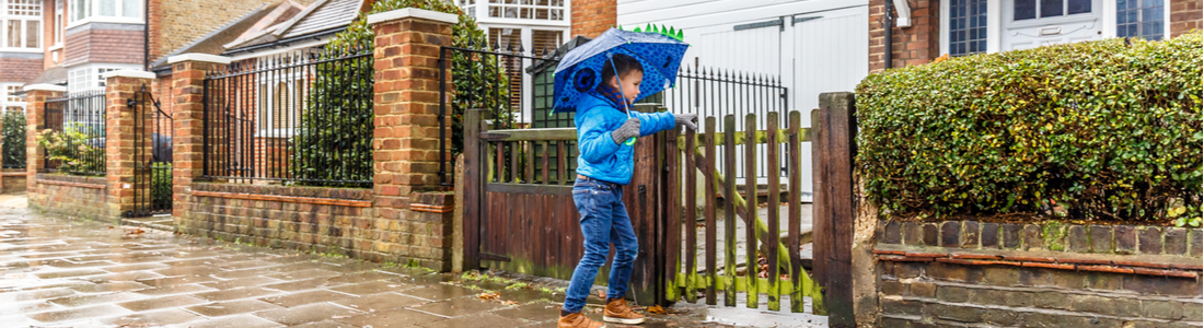A boy with an umbrella opening a gate to a home.